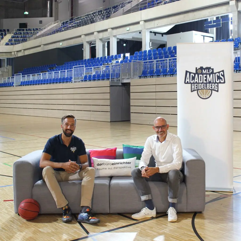 Zwei Männer auf einem Sofa in einer leeren Sporthalle mit Basketball und MLP Academics Heidelberg Banner.