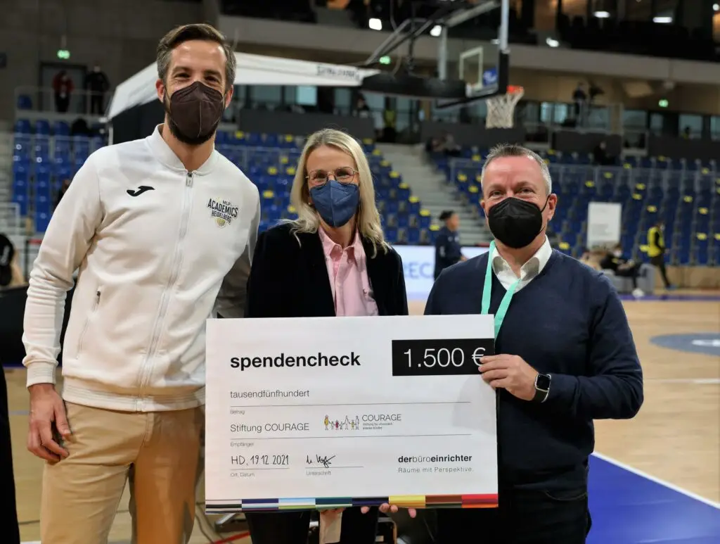 Drei Personen mit Gesichtsmasken halten einen 1.500 € Scheck für Stiftung Courage auf Basketballplatz.