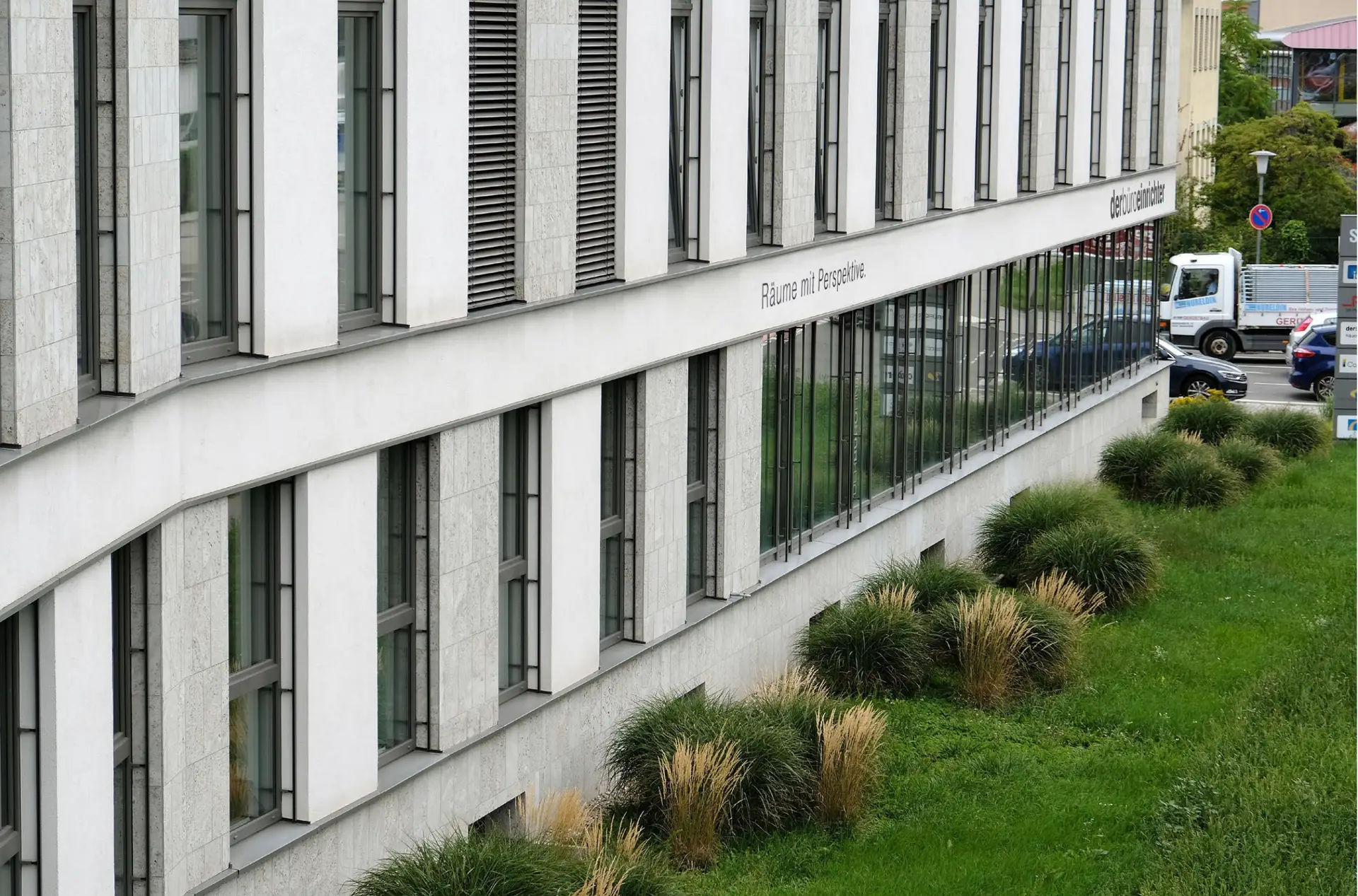 Modernes Bürogebäude mit großen Fenstern, Steinverkleidung und gepflegter Grünfläche davor.