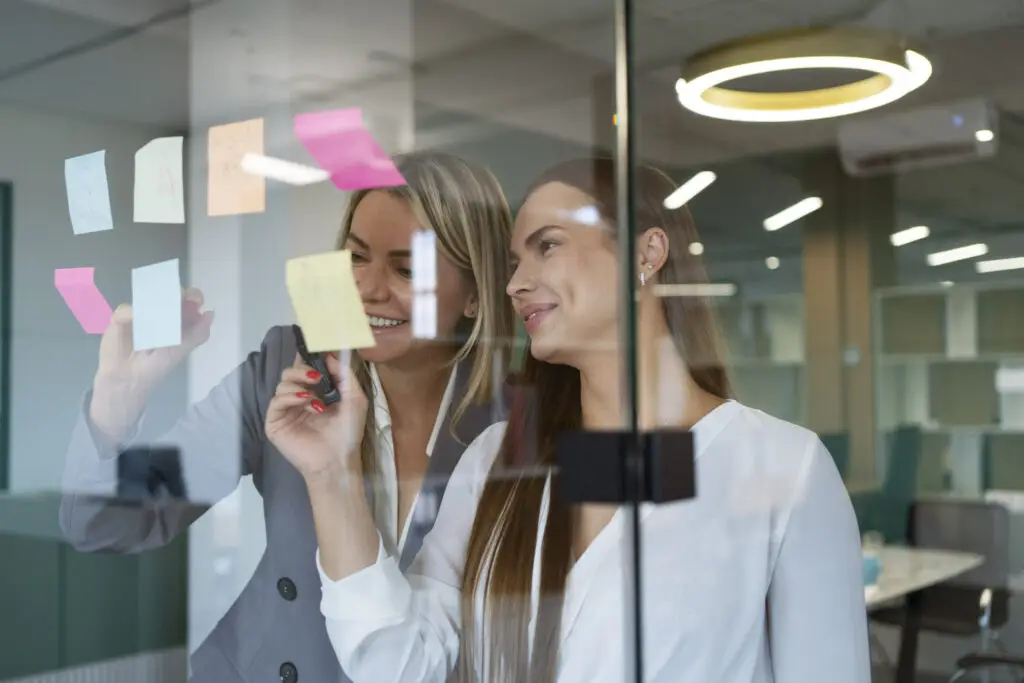 Zwei Frauen arbeiten an einer Glaswand mit Haftnotizen in einem modernen Büro, eine schreibt, die andere sieht zu.