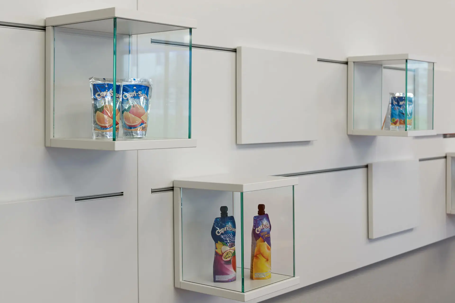 Display mit Capri-Sun-Packungen in gläsernen Regalen an einer weißen Wand.