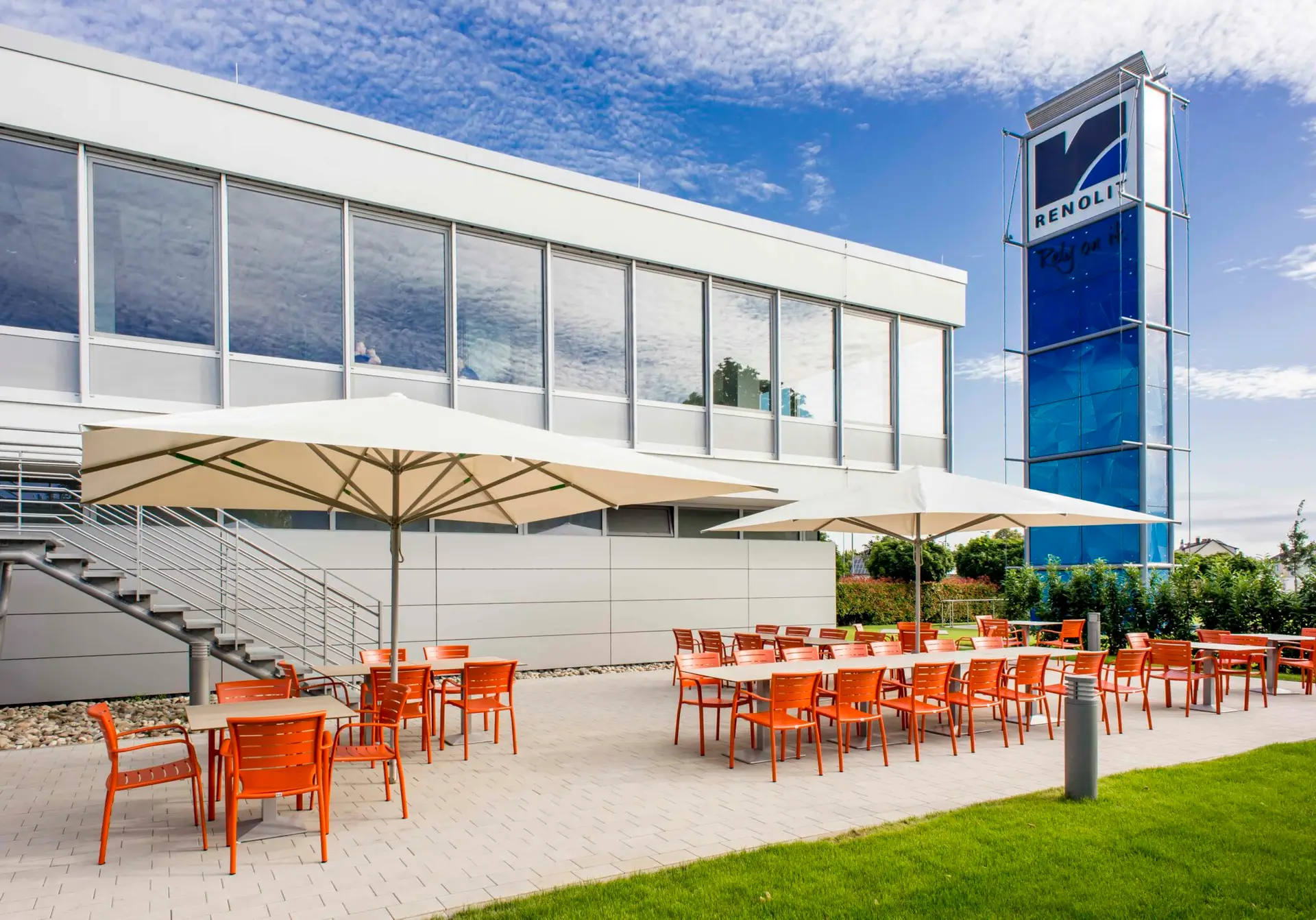 Terrasse eines modernen Gebäudes mit orangenen Möbeln und weißen Sonnenschirmen neben einem blauen Turm.