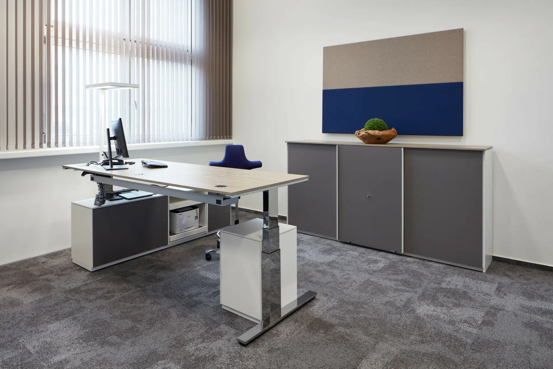 Modernes Büro mit Holztisch, Metallbeinen, Flachbildschirm, Jalousien, grauem Schrank und blauem Stuhl.