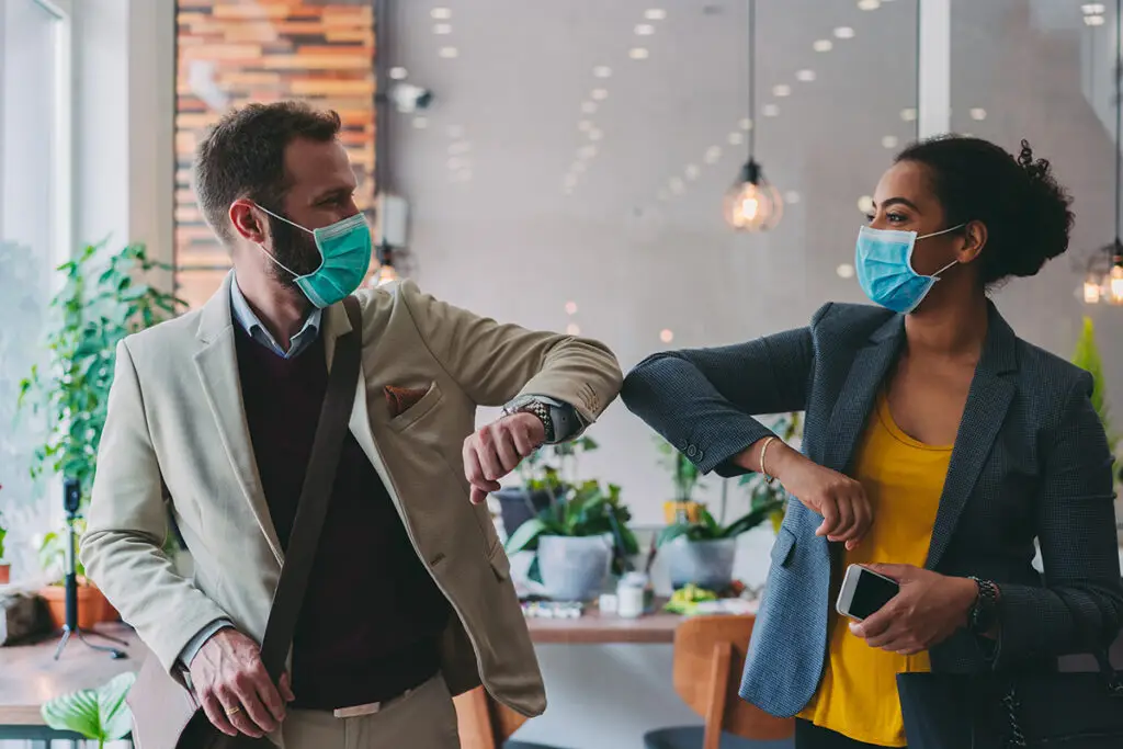 Zwei Personen mit Masken begrüßen sich im modernen, gut beleuchteten Innenraum, Ellbogenstoß.