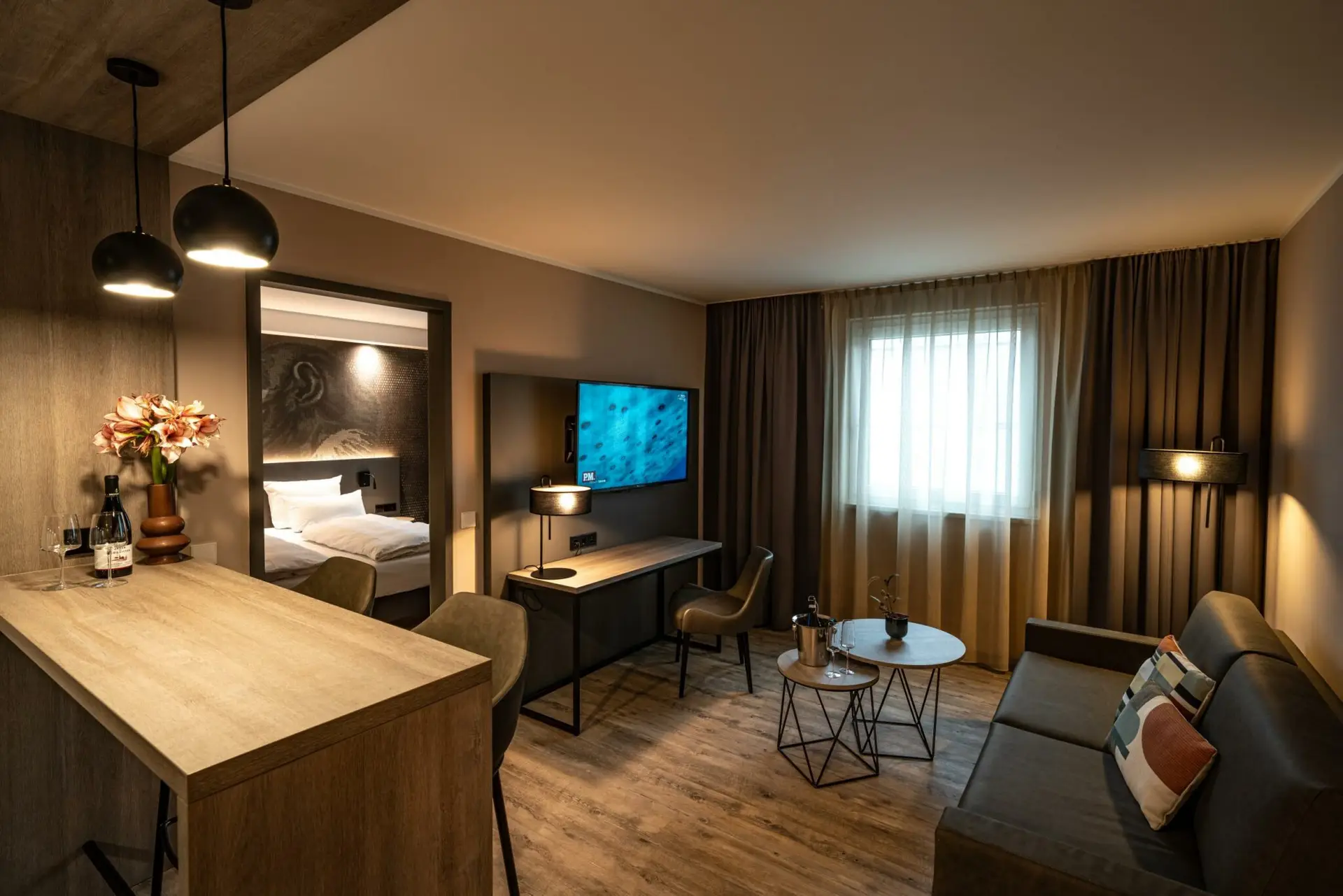 Modernes Hotelzimmer mit Holzboden, Bar, Sofa, Schreibtisch und angrenzendem Schlafzimmer.