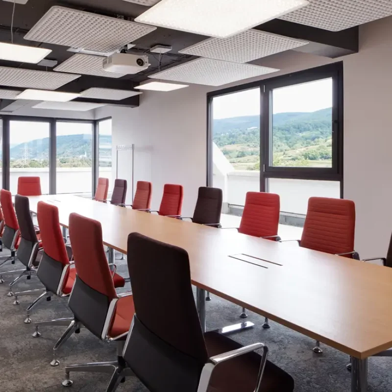 Moderner Konferenzraum mit großem Tisch, rot-braunen Stühlen, Fenstern und LED-Leuchten.