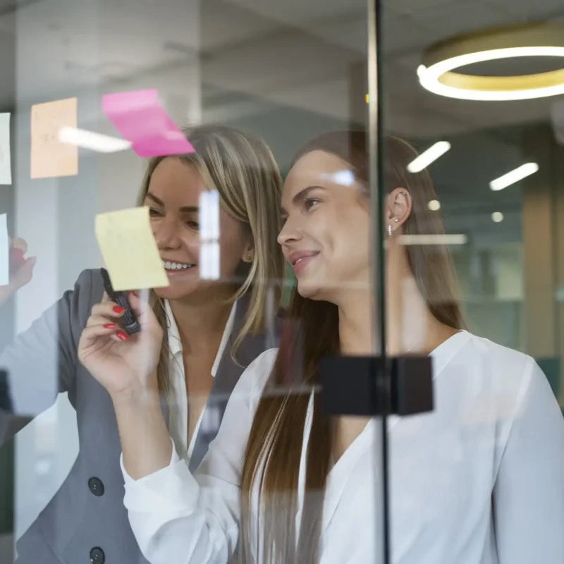 Zwei Frauen arbeiten an einer Glaswand mit Haftnotizen in einem modernen Büro, eine schreibt, die andere sieht zu.